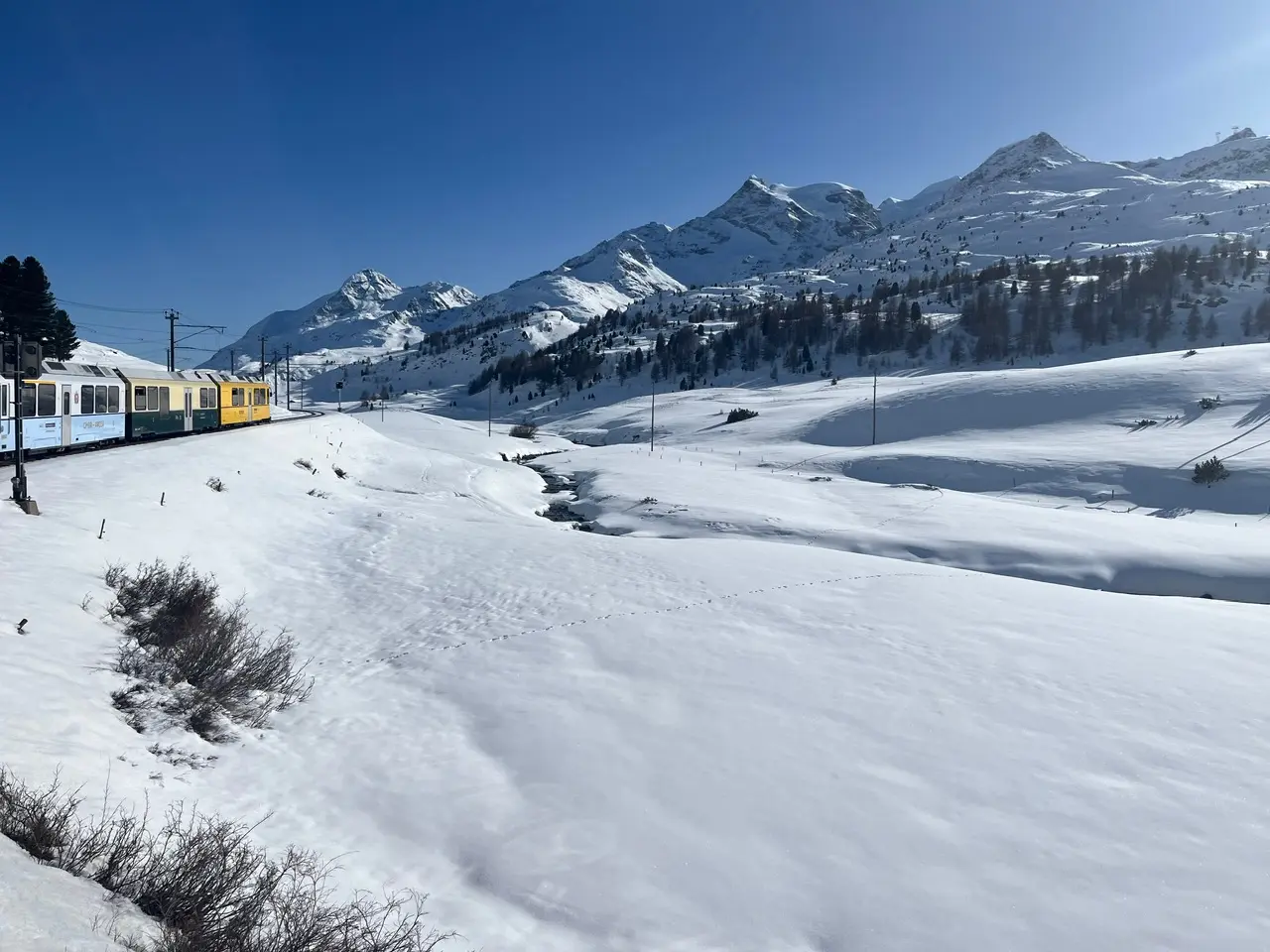 The regional Bernina train visible from the window as it curves through deep snow near the Bernina Pass