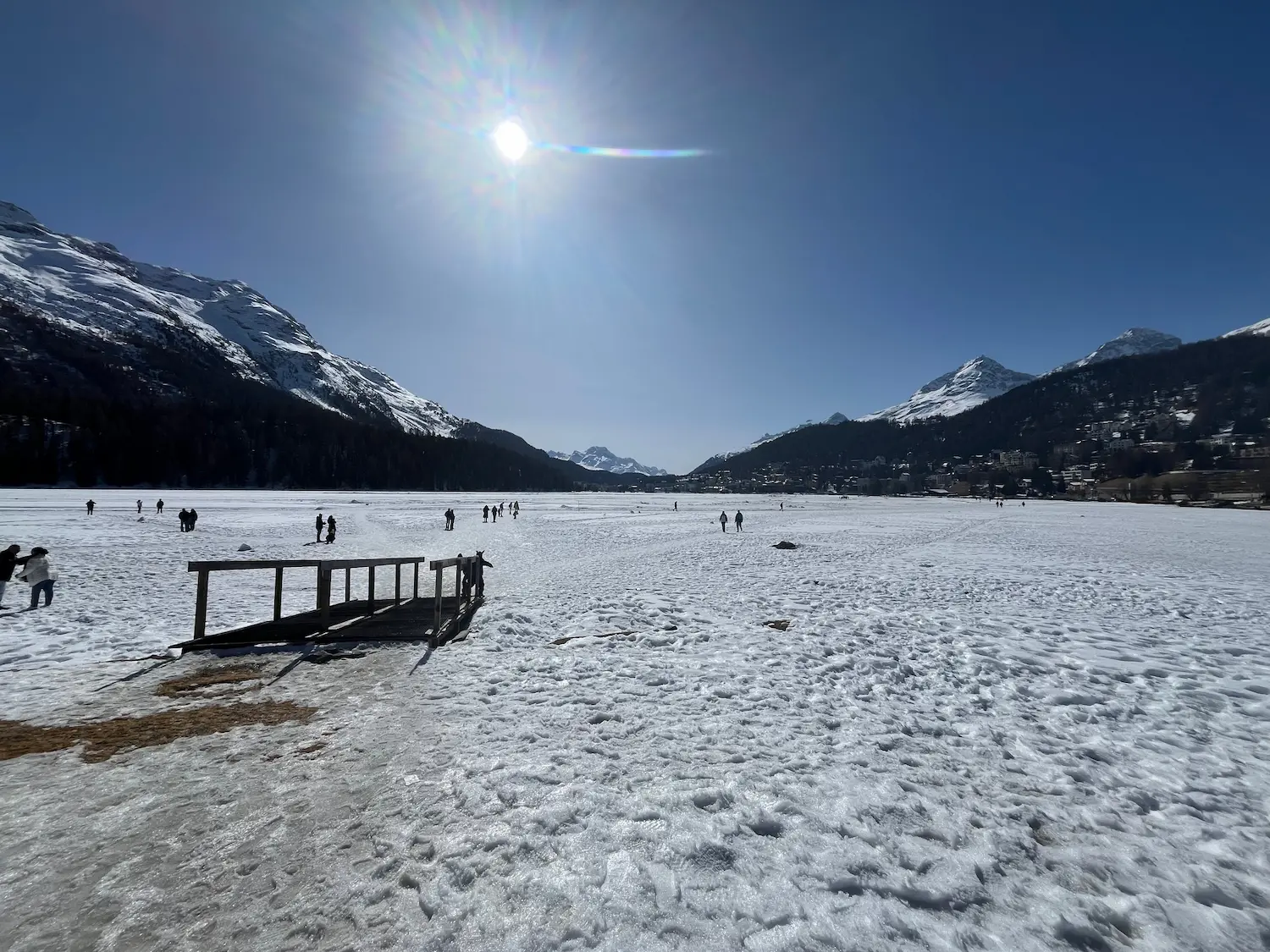 Lake St Moritz frozen solid in winter, with the Alps behind and a wooden jetty in the foreground — people walking across the ice