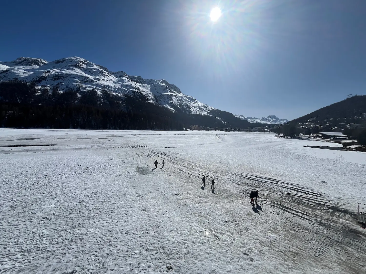 Walkers crossing the frozen surface of Lake St Moritz in bright winter sunshine, St Moritz village visible in the distance