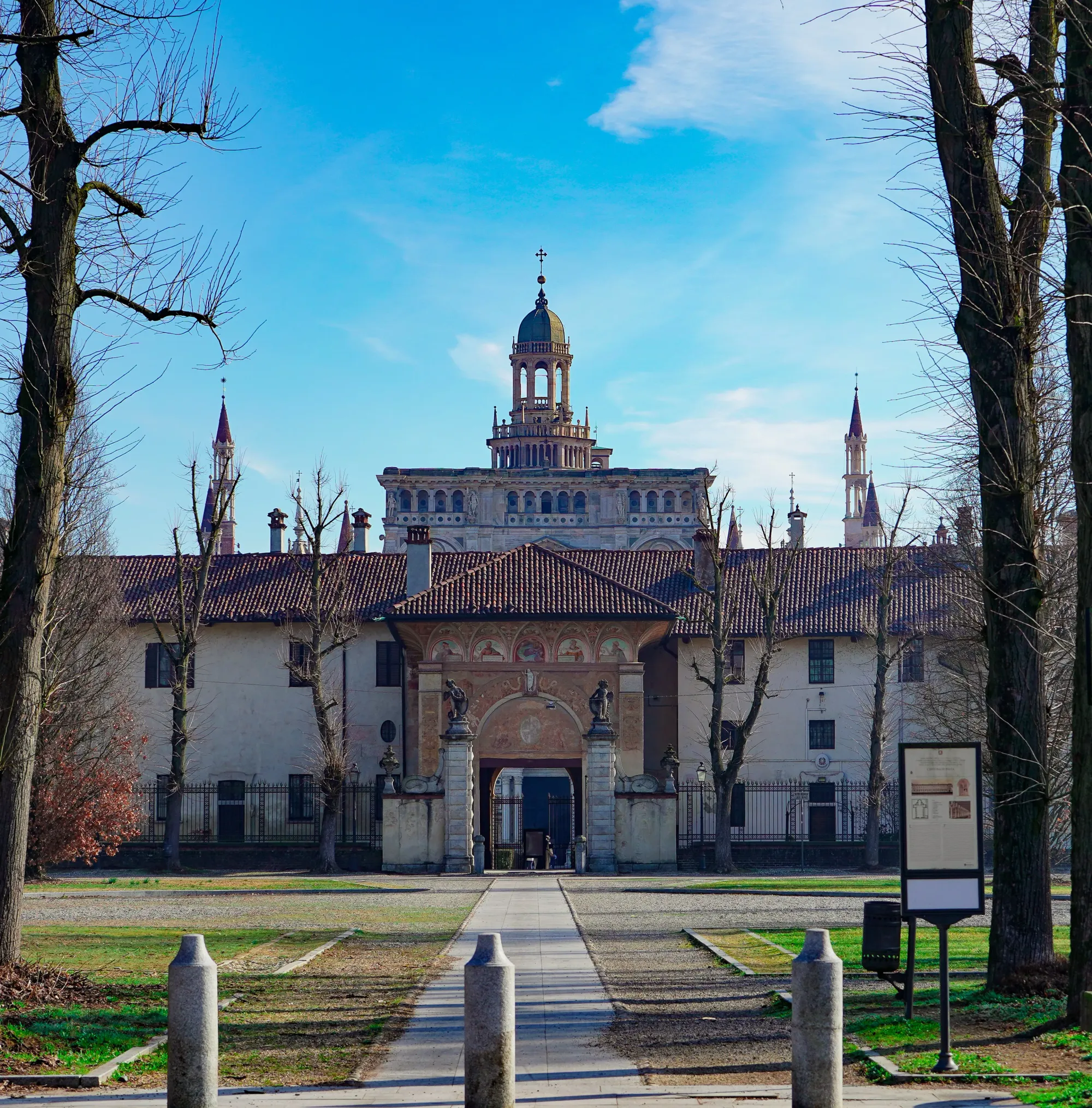 The tree-lined approach path leading to the entrance gate of the Certosa di Pavia, with the ornate tower rising above