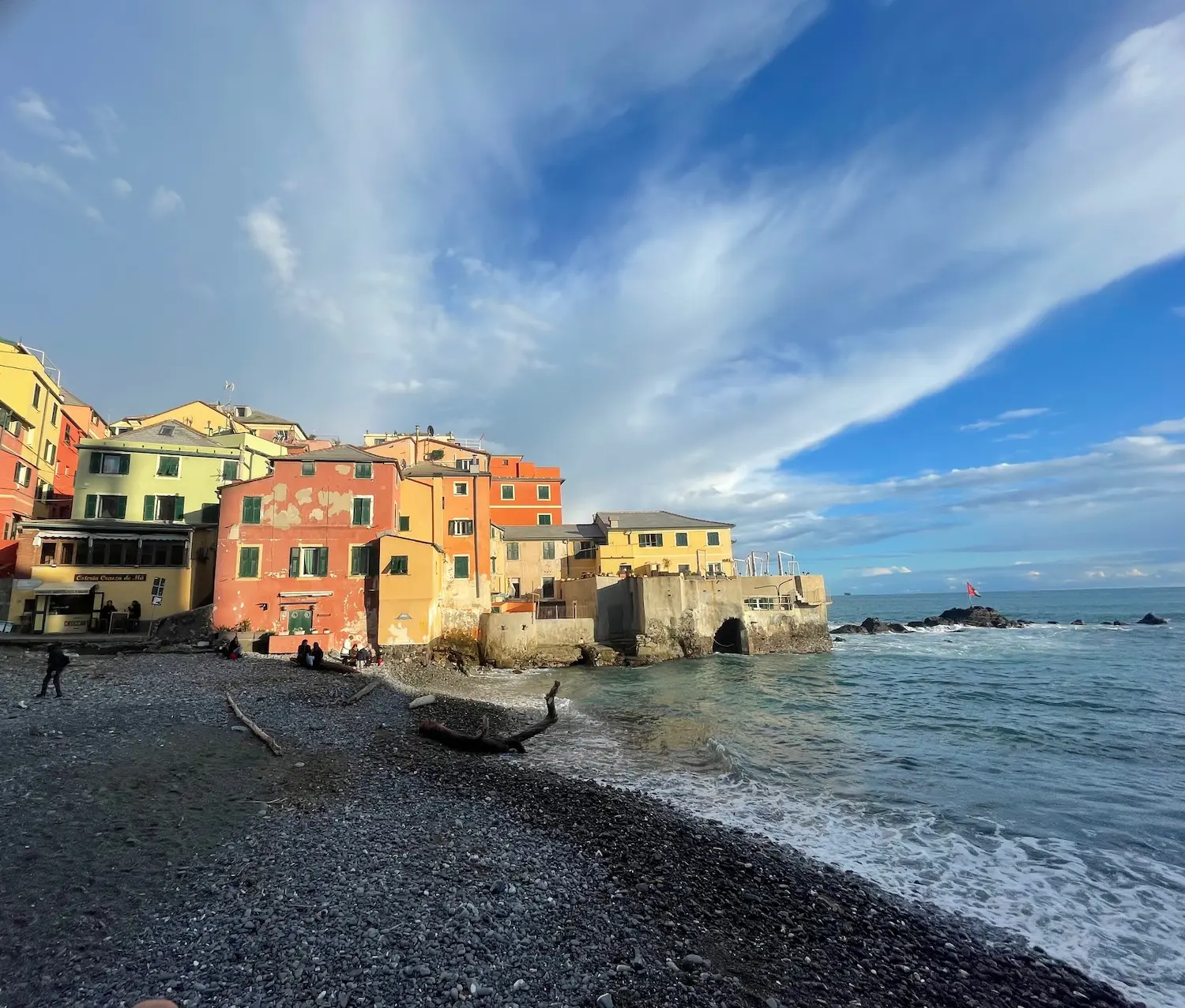 Two figures standing on the rocky breakwater at Boccadasse at dusk, silhouetted against a dramatic sunset sky over the Ligurian Sea