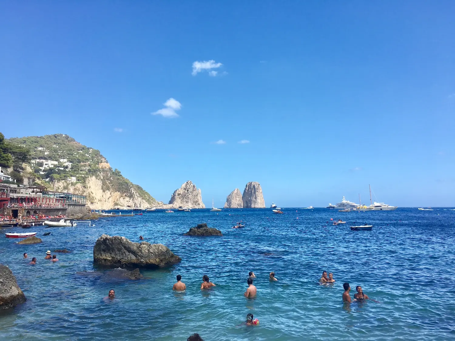 Swimmers in the clear turquoise water at Marina Piccola, Capri, with the iconic Faraglioni rock stacks rising from the sea behind them