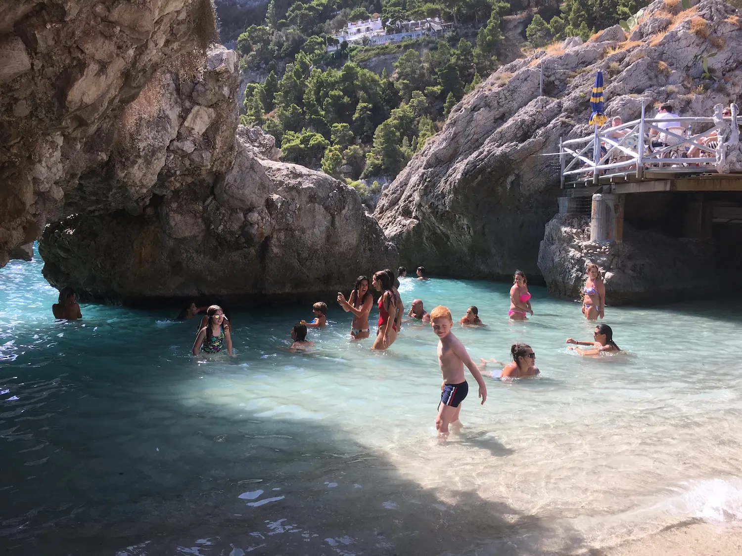 Bathers in a sheltered rocky cove at Marina Piccola, Capri — crystal clear shallow water between limestone cliffs with a beach bar terrace above