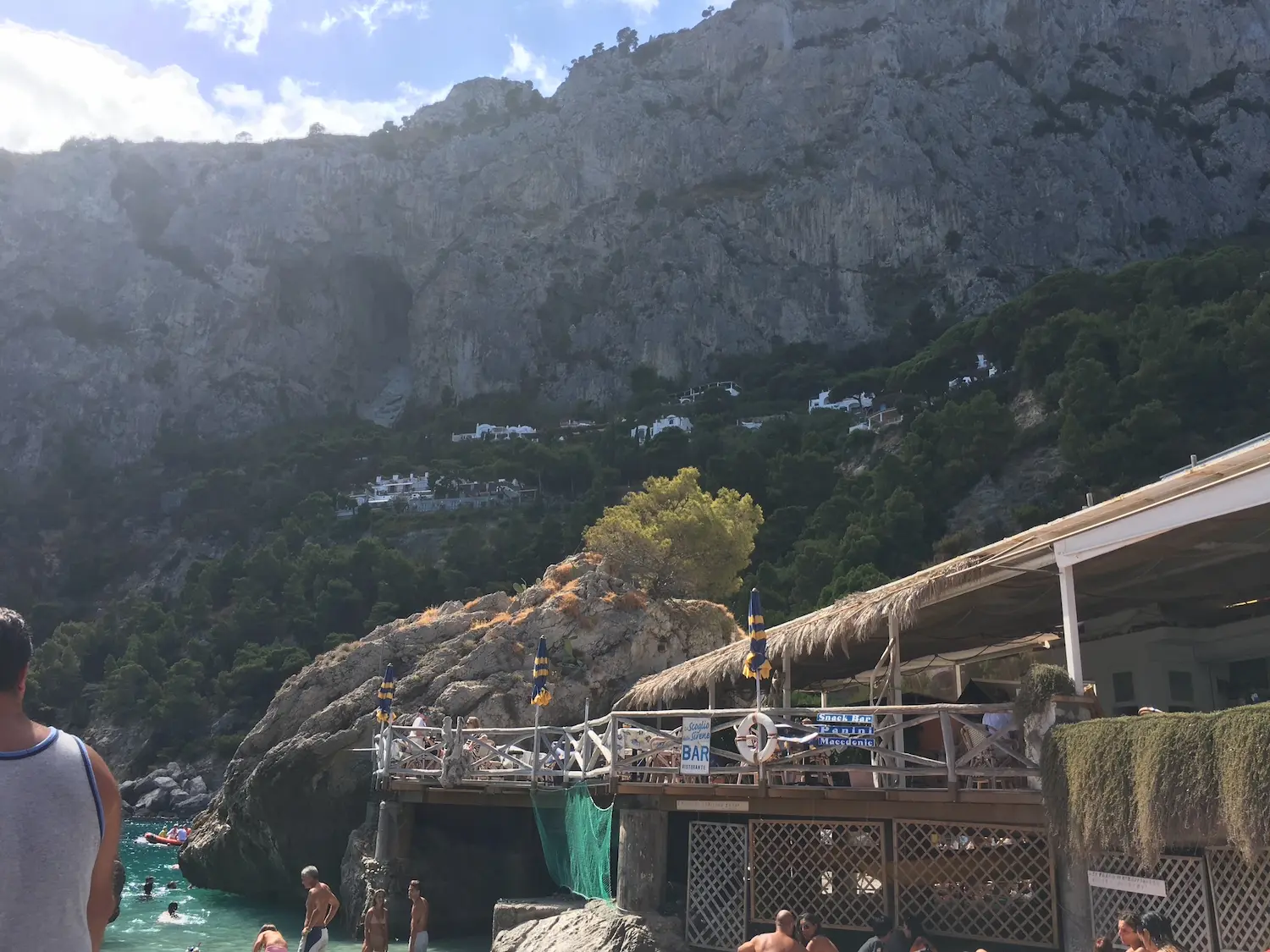 The beach bar and restaurant at Marina Piccola built into the limestone cliff face, with swimmers below and the wooded hillside of Capri rising steeply behind